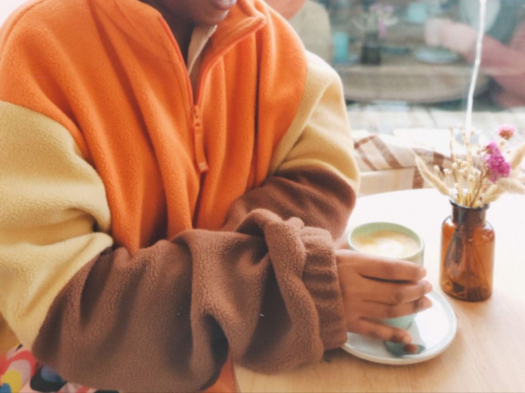 Close-up of a person in a colorful fleece holding a fresh latte at a cafe table.